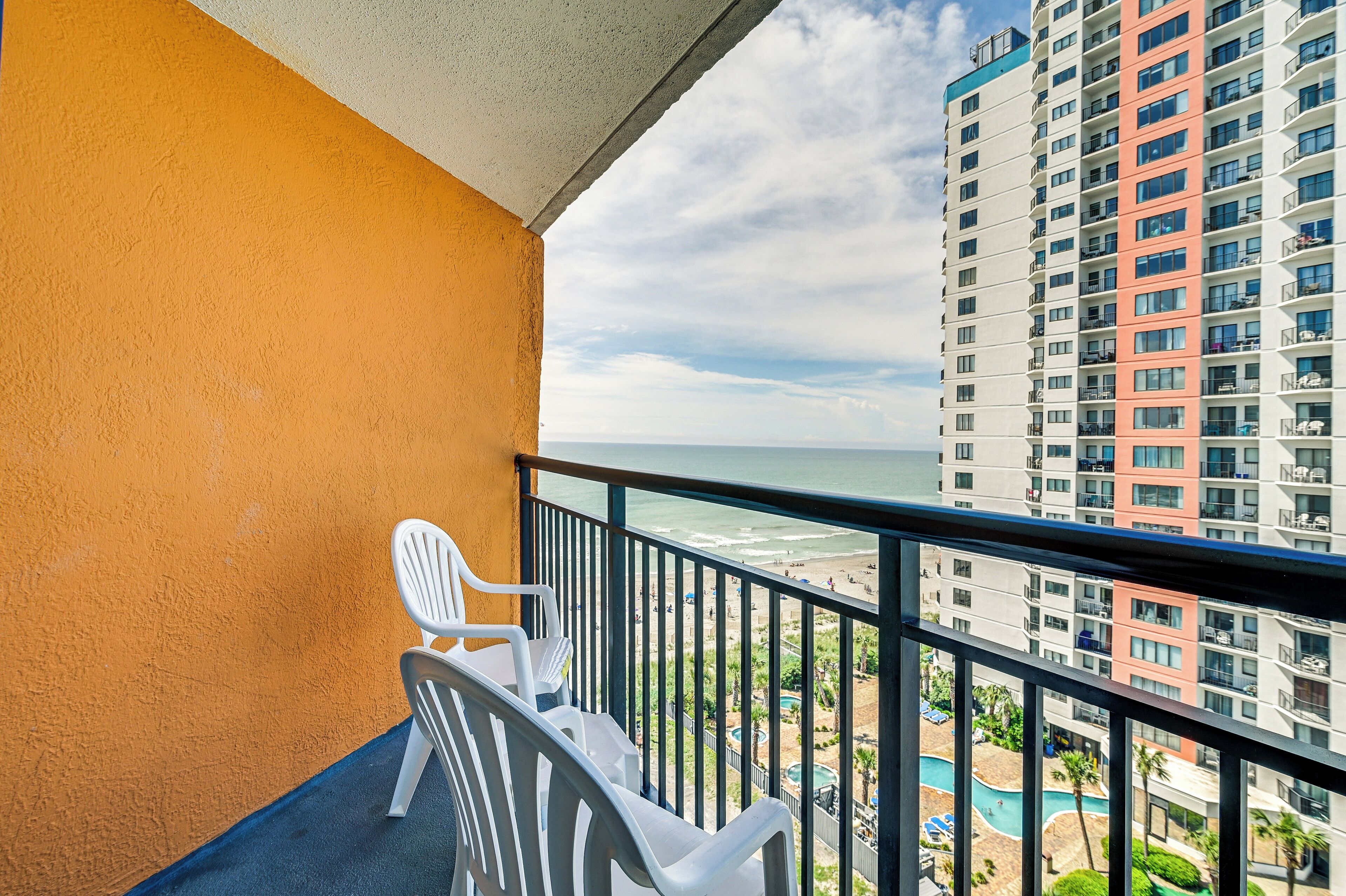 Un balcon avec des chaises blanches, donnant sur une plage et un immeuble de grande hauteur également doté de balcons.