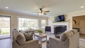 A spacious living room with a fireplace, a ceiling fan, and large windows.