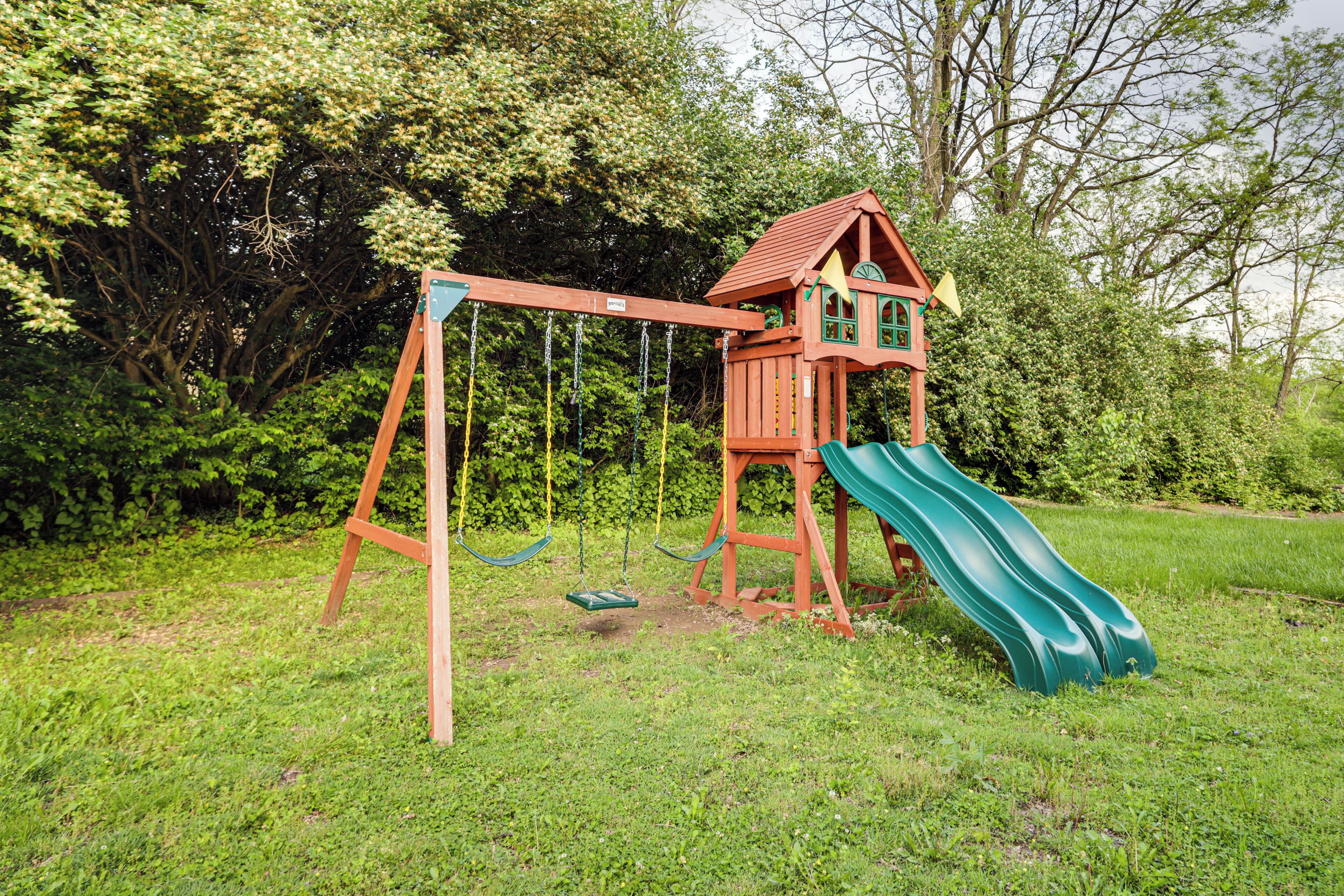A wooden playhouse with a slide and swings in a grassy area.
