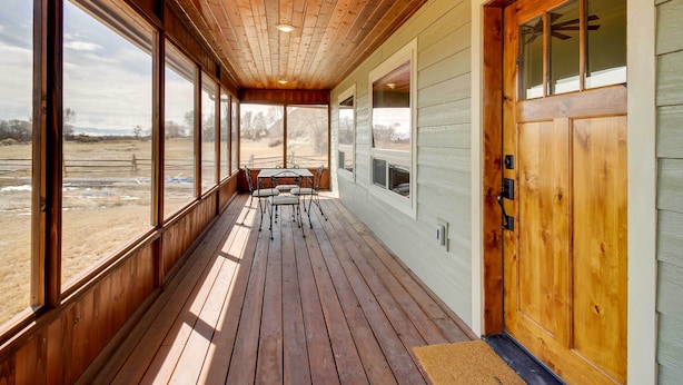 A covered porch with wooden flooring, a table with chairs, and large windows offering views of a rural landscape. - Beautiful Jefferson River Cabin w/ Mountain Views! (Twin Bridges)
