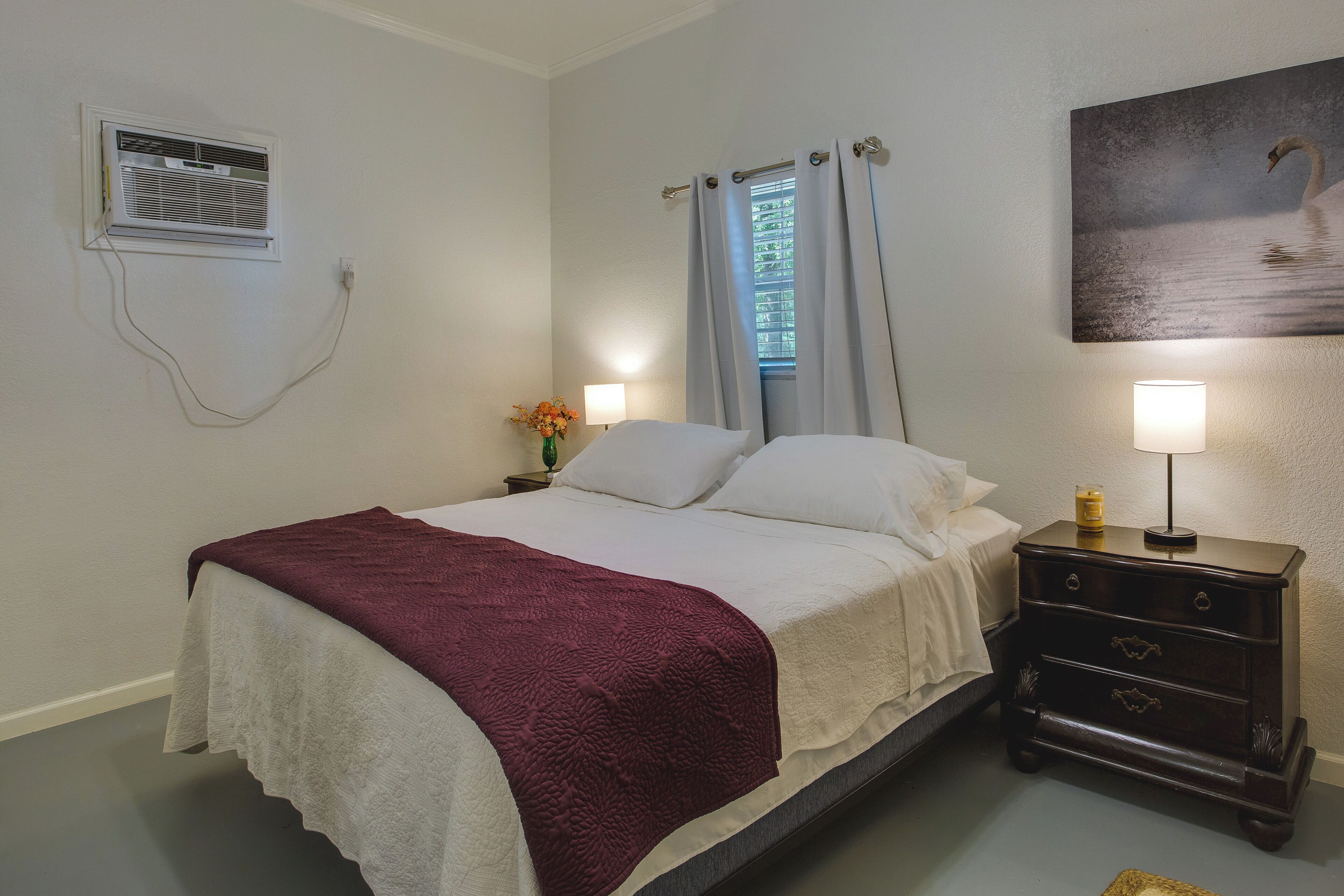 Un dormitorio con cama, mesita de noche, lámpara, ventana con cortinas y aire acondicionado instalado en la pared.