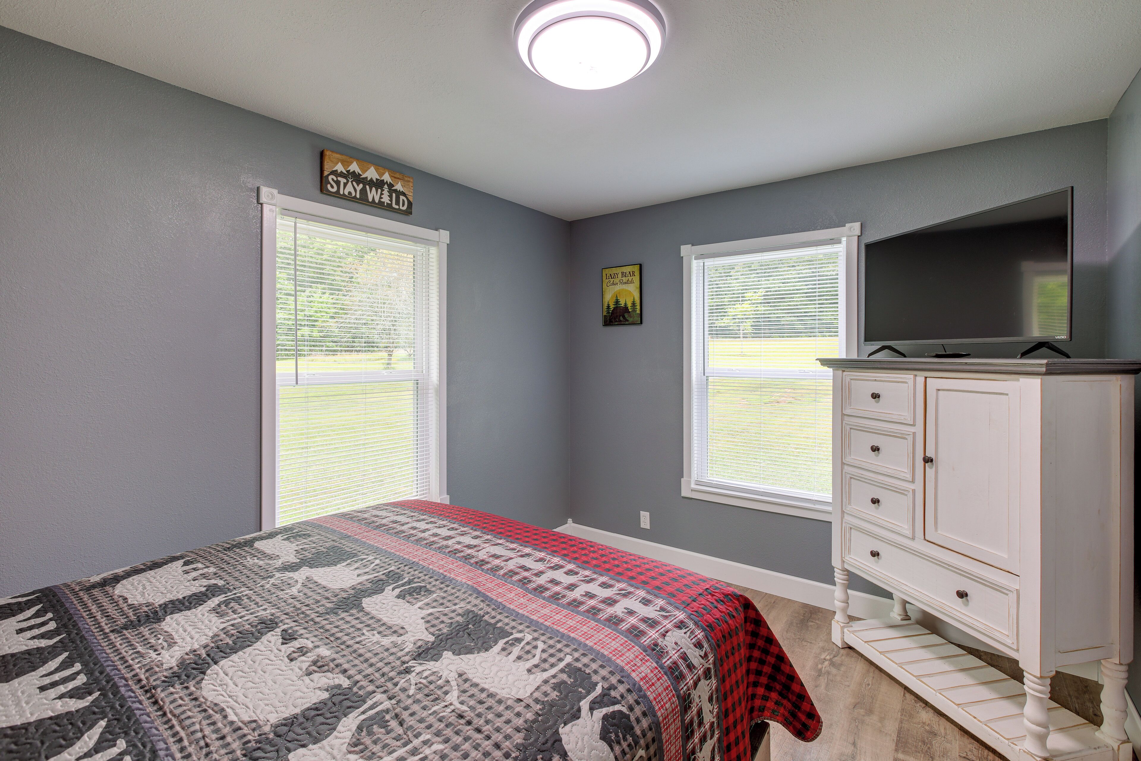A bedroom with a bed, dresser, and TV. There are two windows with blinds, a ceiling light, and a sign on the wall.