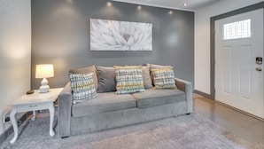 A living room with a grey sofa, patterned cushions, a white side table with a lamp, and a large floral artwork on the wall.
