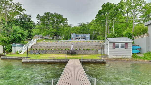 Une jetée en bois s’étendant sur une étendue d’eau calme, entourée d’une végétation luxuriante et bordée d’un petit bâtiment doté d’un mur en pierre.