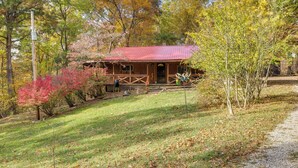 A rustic cabin with a red roof, surrounded by autumn foliage and a gravel path. - French Lick Getaway w/ Deck, Grill & Fire Pit! (French Lick)