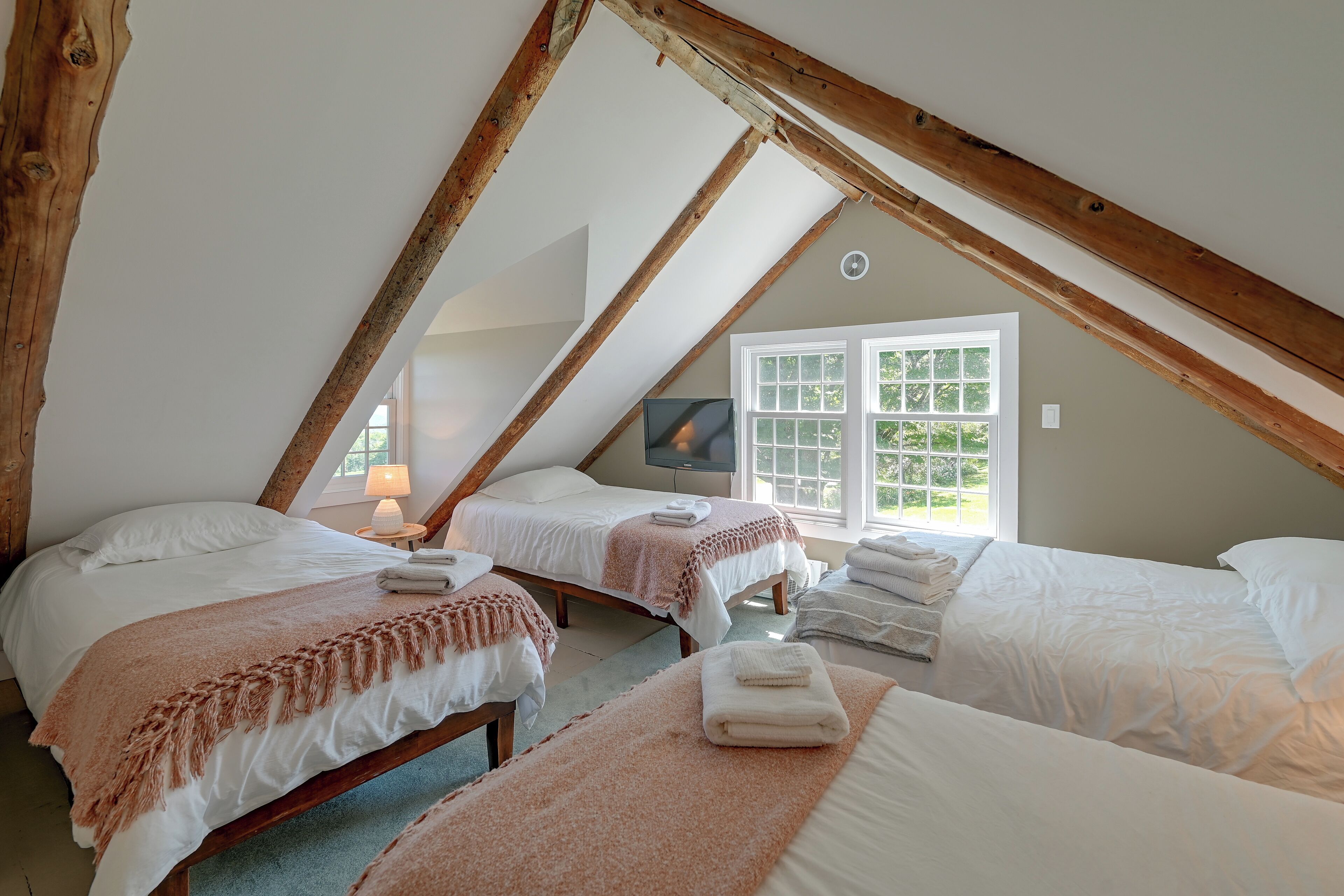 A cozy attic bedroom with two beds, a television, and a window with a view of greenery.