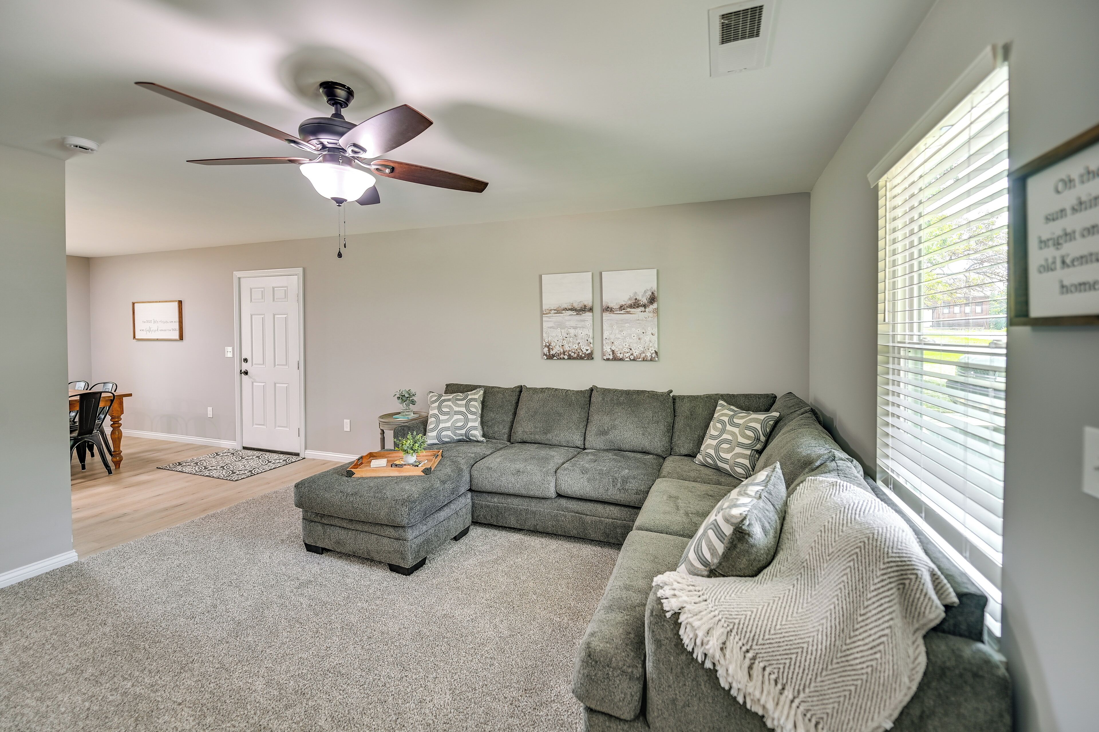 A living room with a sectional sofa, ottoman, ceiling fan, and artwork on the wall.