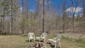 An outdoor seating area with four white chairs arranged around a fire pit in a forested setting. - Near Waterville: Cozy Midcoast Vacation Rental! (Albion)