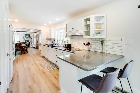 A modern kitchen with white cabinetry, a central island with bar stools, and a dining area with wooden chairs and a table. - Pickleball Court & Fire Pit: Milton Cabin! (Milton)