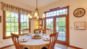 A dining room with a round table, six wooden chairs, a chandelier, and large windows with a view of greenery.