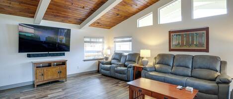 A spacious living room with a wooden ceiling, a large flat-screen TV, leather sofas, a wooden coffee table, and a painting on the wall.