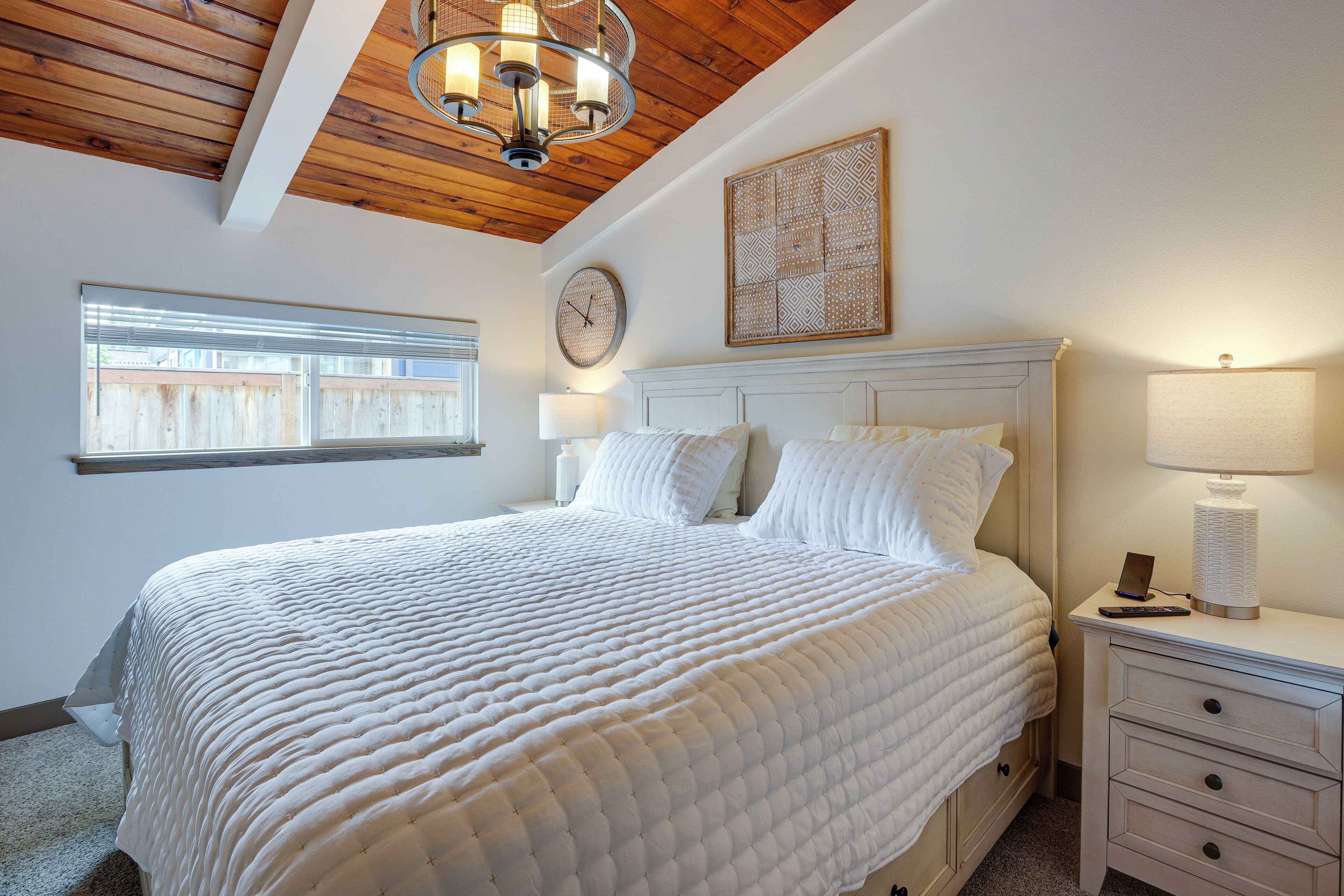 Een slaapkamer met een groot bed, een houten plafond, een wandklok, een nachtkastje met een lamp en een raam met zonwering.