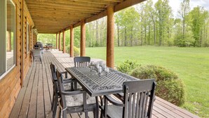 A covered outdoor patio with a wooden table and chairs, a grill, and a view of a grassy area with trees.
