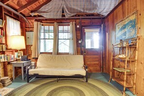 Ein gemütliches Blockhaus mit Bett, Schreibtisch mit Lampe, Bücherregal und großem Fenster mit Vorhängen.