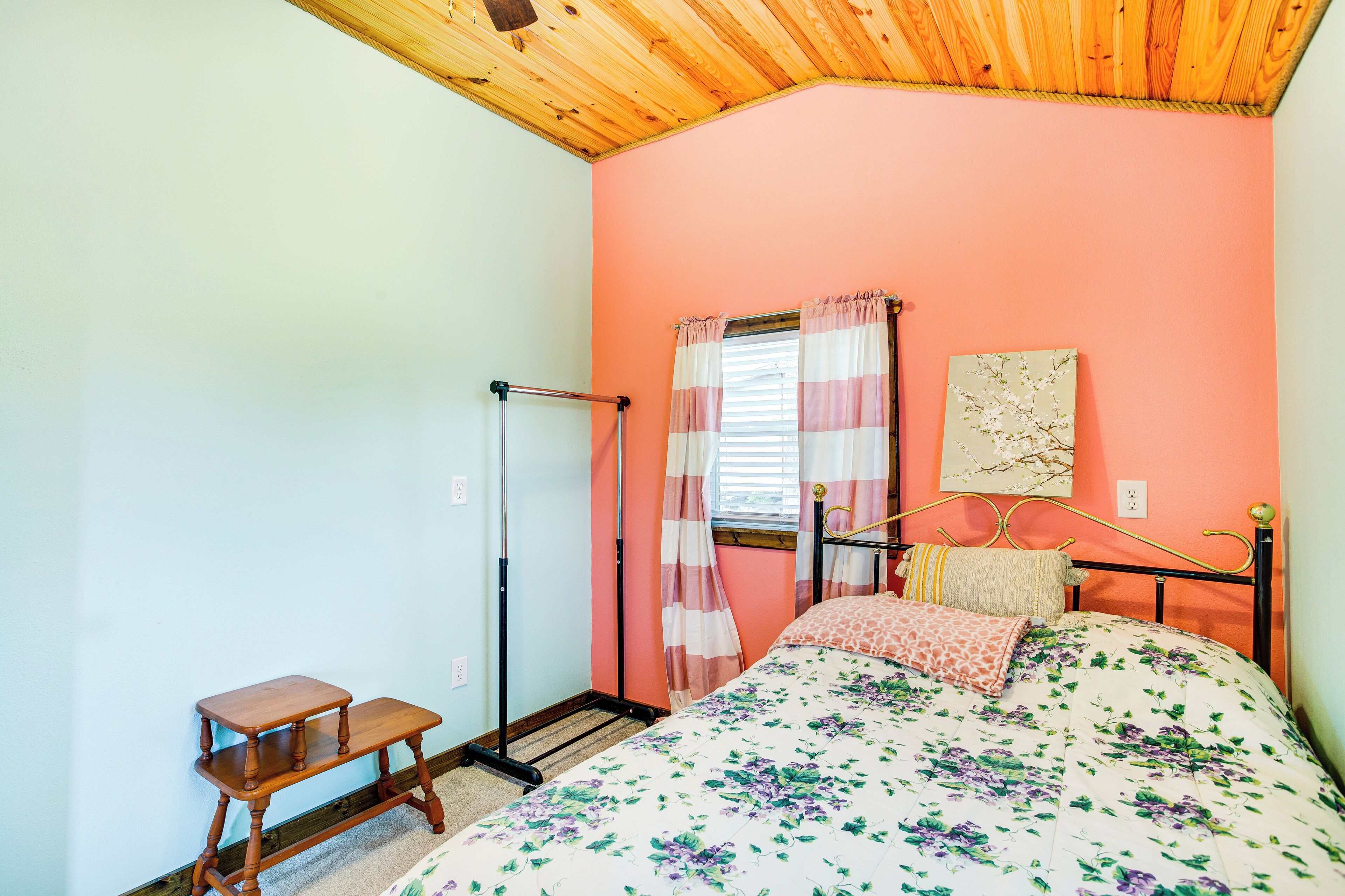 A bedroom with a bed, a wooden stool, a window with curtains, and a wall-mounted artwork.