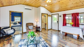 A room with a wooden ceiling, a bed, a chair, a table with a vase of flowers, and a window with red curtains. - Northeast Texas Vacation Rental ~ 6 Mi to Commerce (Commerce)