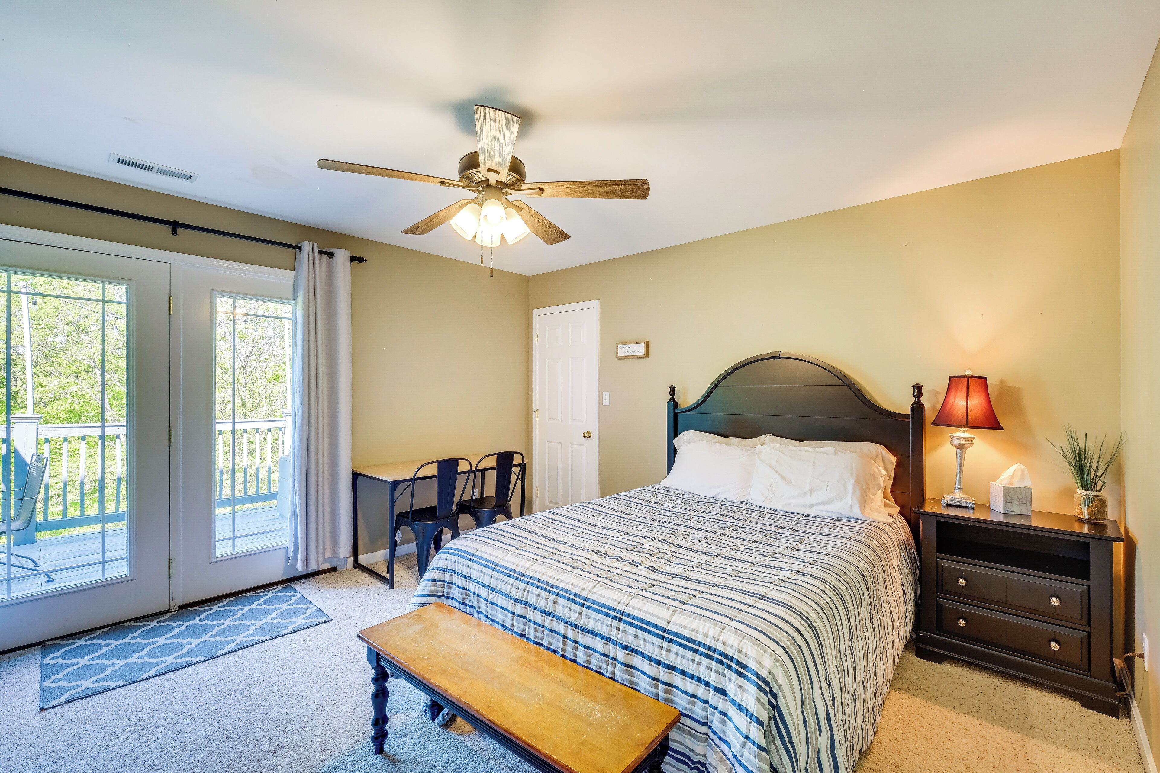 A bedroom with a bed, a nightstand, a bench, a ceiling fan, and a view of the outdoors through a sliding door.