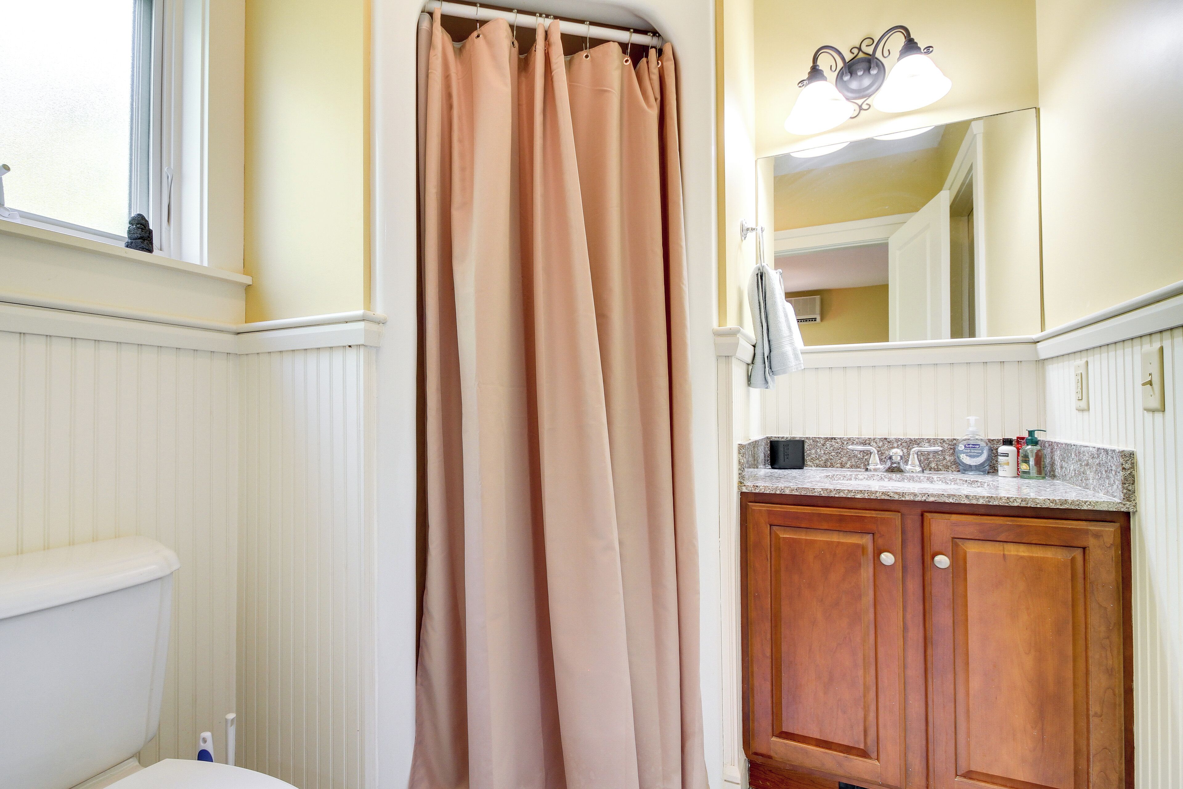 Une salle de bain comprenant des toilettes, un rideau de douche, un meuble-lavabo avec un miroir et un luminaire placé au-dessus du miroir.