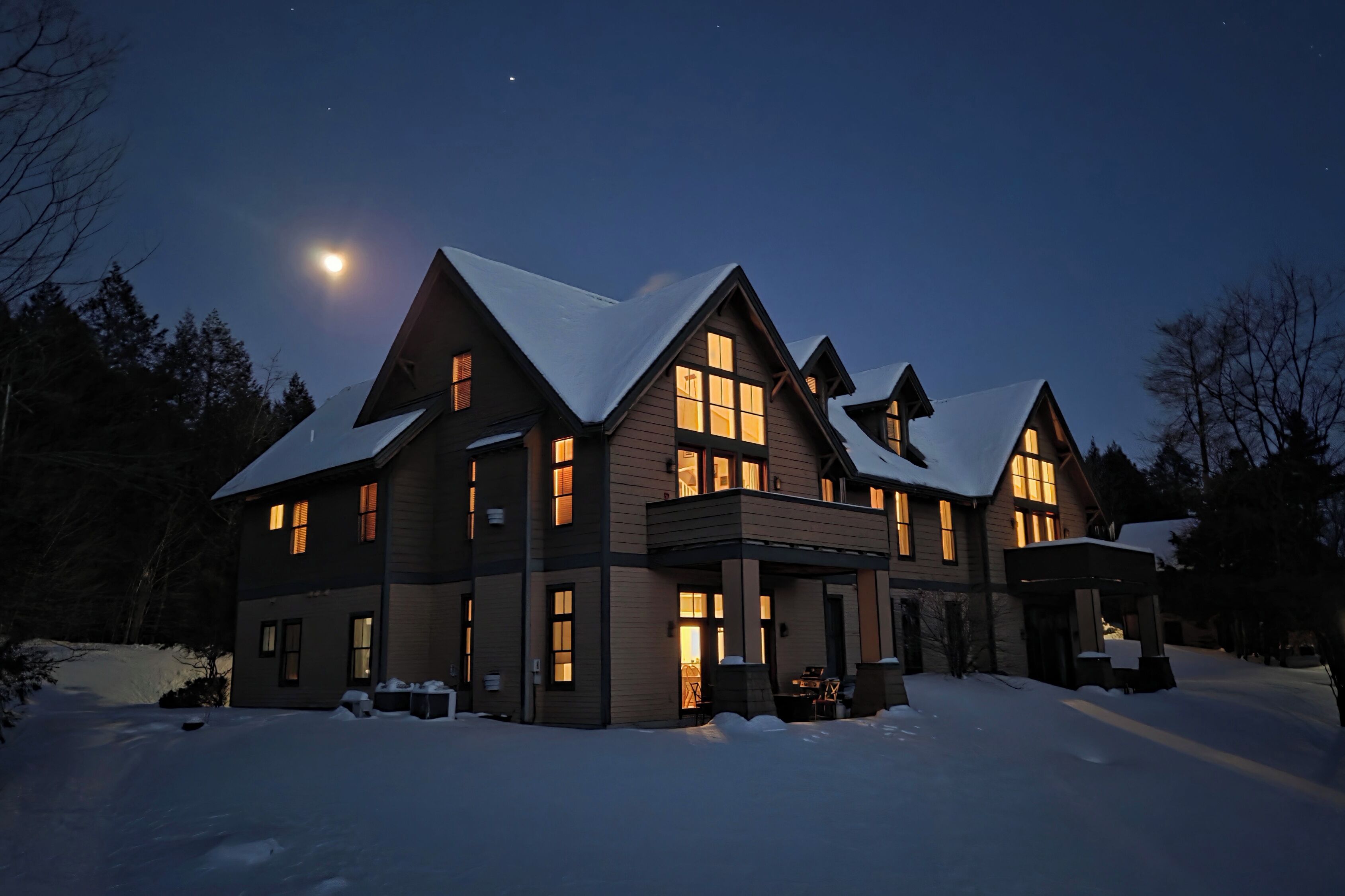 A large, multi-story house with a snow-covered roof and multiple windows, surrounded by trees.