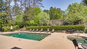 Une piscine entourée d’arbres et d’un mur de pierre, avec des chaises longues et des tables à proximité.