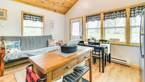 A cozy living area with a wooden ceiling, a gray sofa with a map pillow, a wooden dining table with chairs, and a kitchen area with a refrigerator. - Cozy New York Abode - Porch, Near Fishing & Hiking (Jordan)