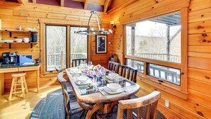 A wooden cabin interior with a dining table set for six, a kitchen area with a coffee maker, and large windows offering views of the outdoors. - Berkshire Hills Nature Lovers' Log Home Retreat (Windsor)