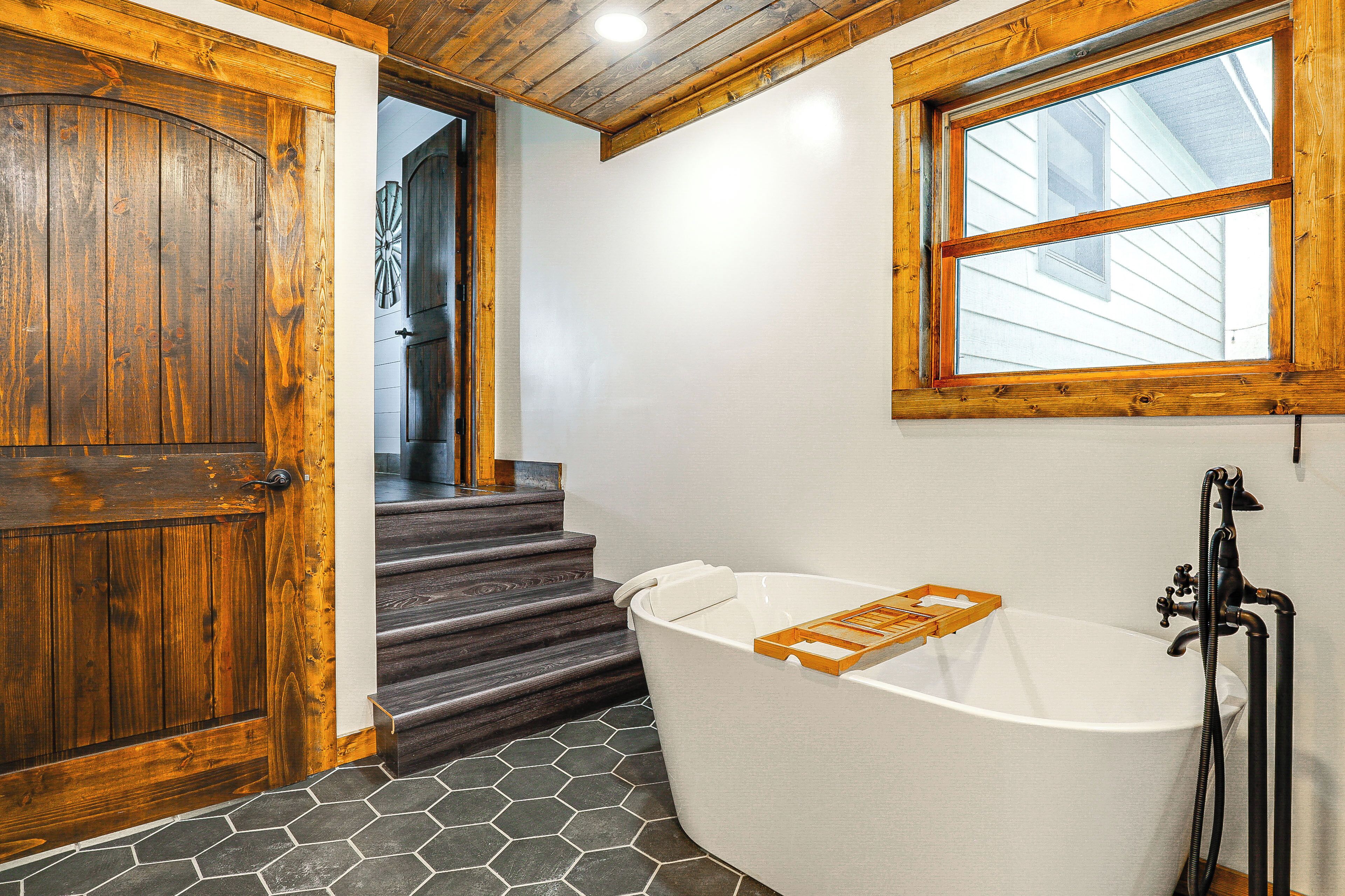 A bathroom with a wooden door, a white bathtub with a wooden tray, and a window above the tub.