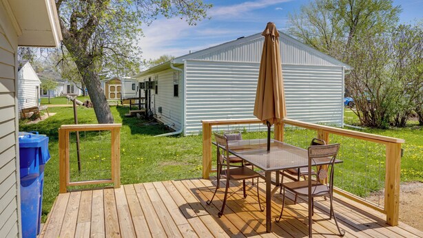 A wooden deck with a table and chairs, a trash bin, and a fenced area with a small building in the background. - Vacation Rental Near South Dakota State University (Aurora)