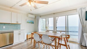 A spacious kitchen with a wooden dining table and chairs, a ceiling fan, and a view of the ocean through large windows. - Beachy Palms Cottage on Lake Huron w/ Hot Tub! (Palms)