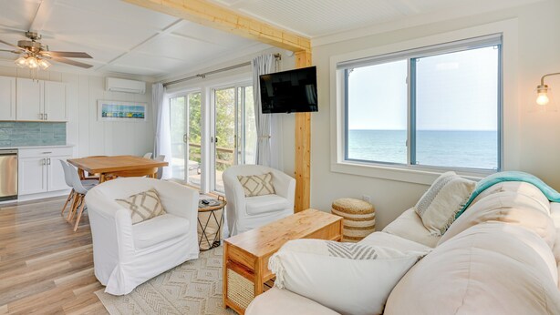 A spacious living area with a wooden ceiling, a large window with a view of the sea, a flat-screen TV mounted on the wall, and a cozy seating arrangement including a sofa and chairs. - Beachy Palms Cottage on Lake Huron w/ Hot Tub! (Palms)