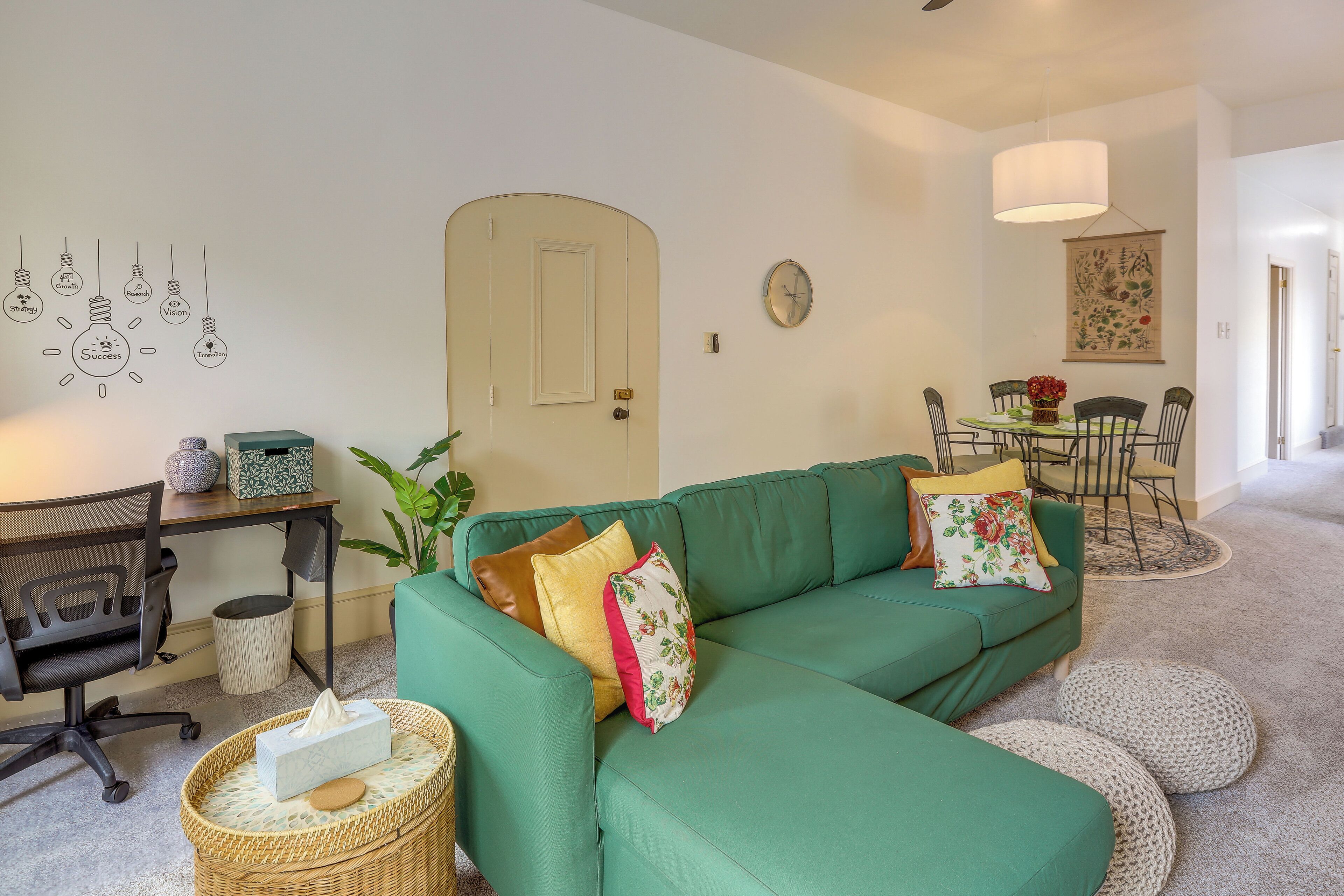 Un salon avec un canapé vert, une table à manger avec des chaises, une horloge murale et un mur décoré de dessins d’ampoules.