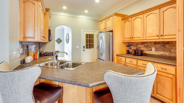 A kitchen with wooden cabinets, granite countertops, and a built-in island with bar stools. - Muscle Shoals Home < 3 Mi to Recording Studios! (Muscle Shoals)