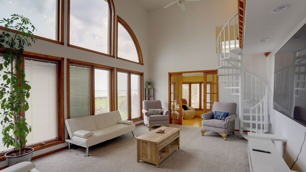 A spacious living room with a white sofa, a wooden coffee table, and two armchairs. There are large windows with wooden frames, a ceiling fan, and a staircase leading to an upper level.
