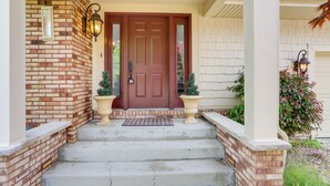 A brick exterior with a red door, two potted plants, and a white porch with steps leading to the door.