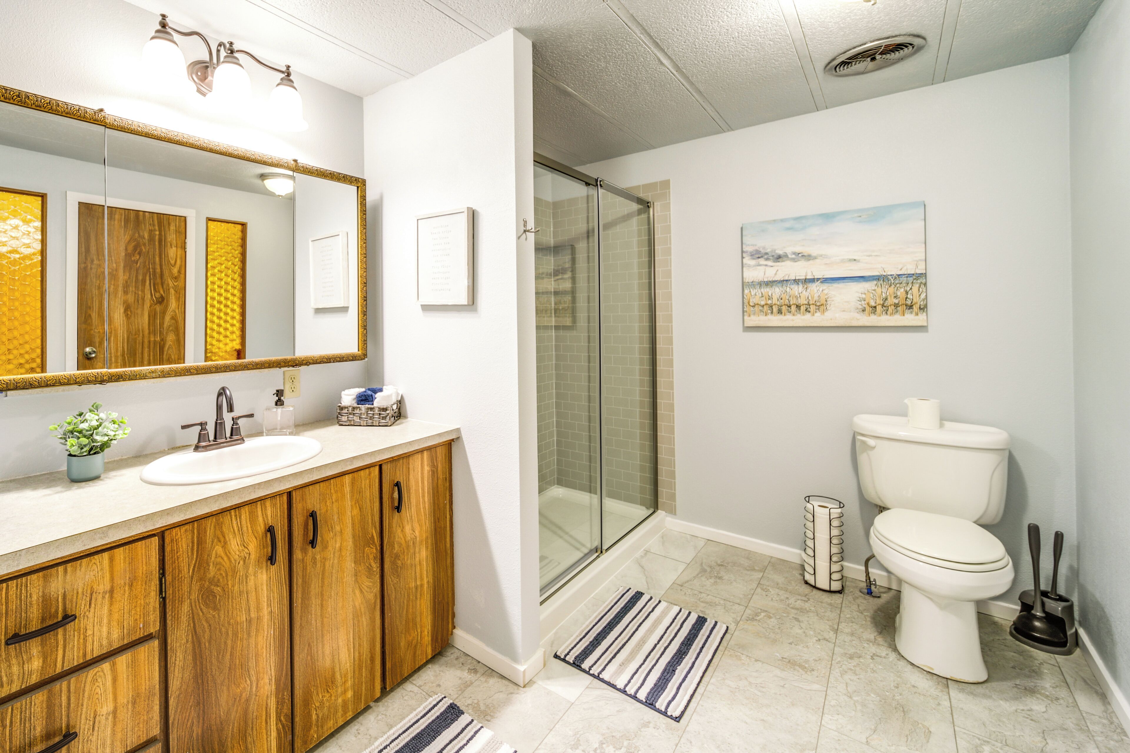 A bathroom with a wooden vanity, a white sink, a glass shower enclosure, a toilet, and a framed landscape painting on the wall.