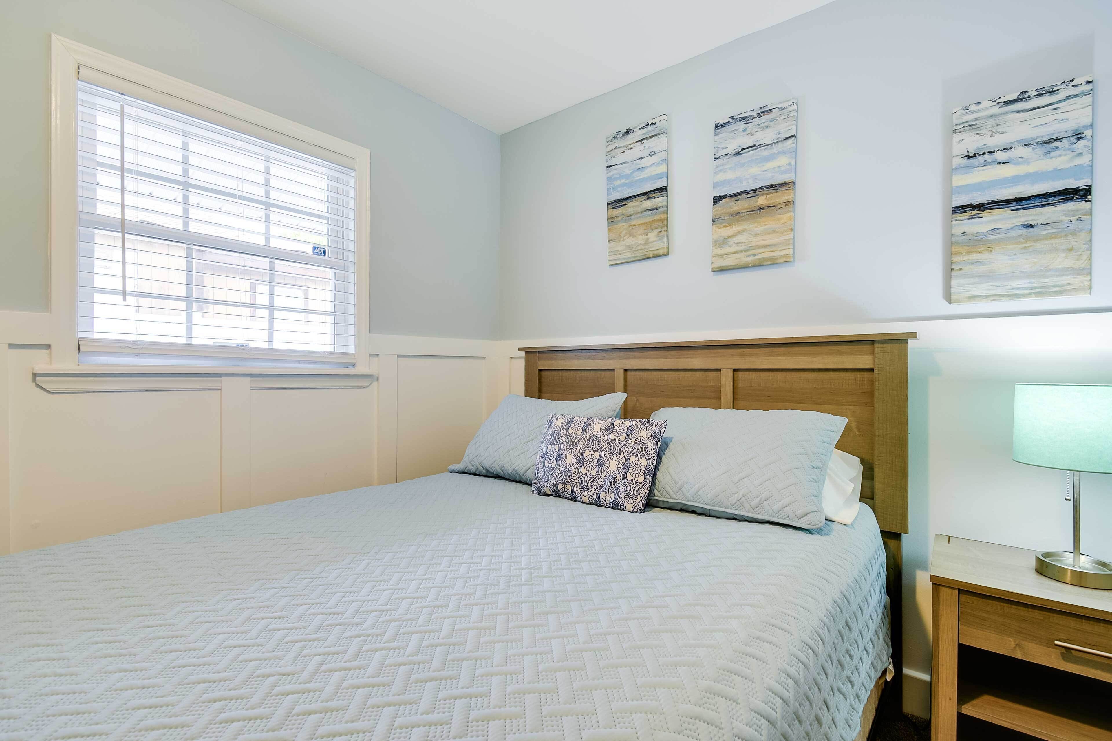 A bedroom with a bed, a nightstand, a window with blinds, and three wall-mounted paintings.