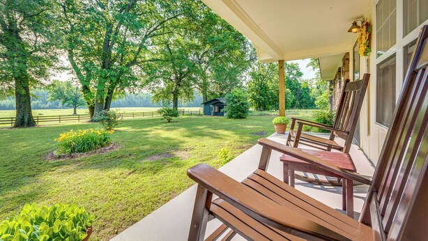 A covered porch with wooden benches and a view of a grassy yard with trees and a small building in the distance. - Delaware Vacation Rental: Walk to Lake Dardanelle! (Delaware)