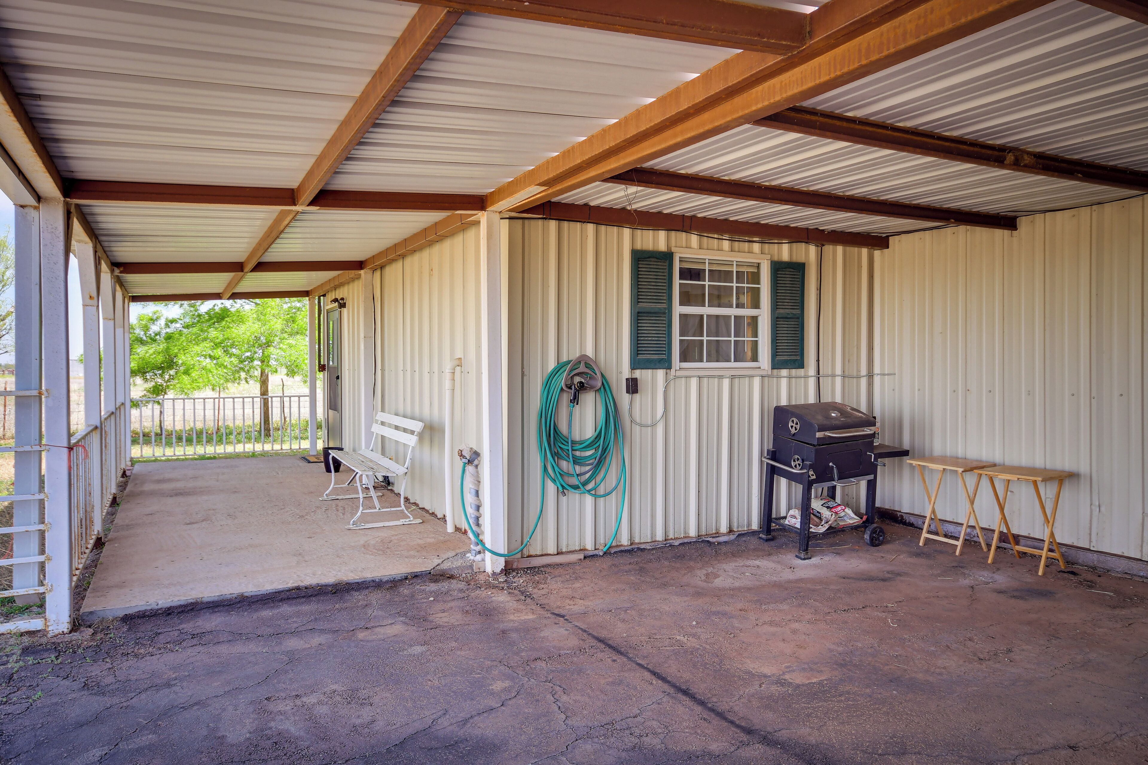 A covered outdoor area with a concrete floor, a metal roof, and a corrugated metal wall. There is a bench, a grill, and a table.
