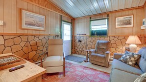 A cozy room with wooden walls, a stone accent wall, and a fireplace. There are two armchairs and a sofa with a patterned pillow. A remote control is on a wooden table. - Stargaze, Hike & Kayak: Cabin Near Copper Breaks! (Crowell)