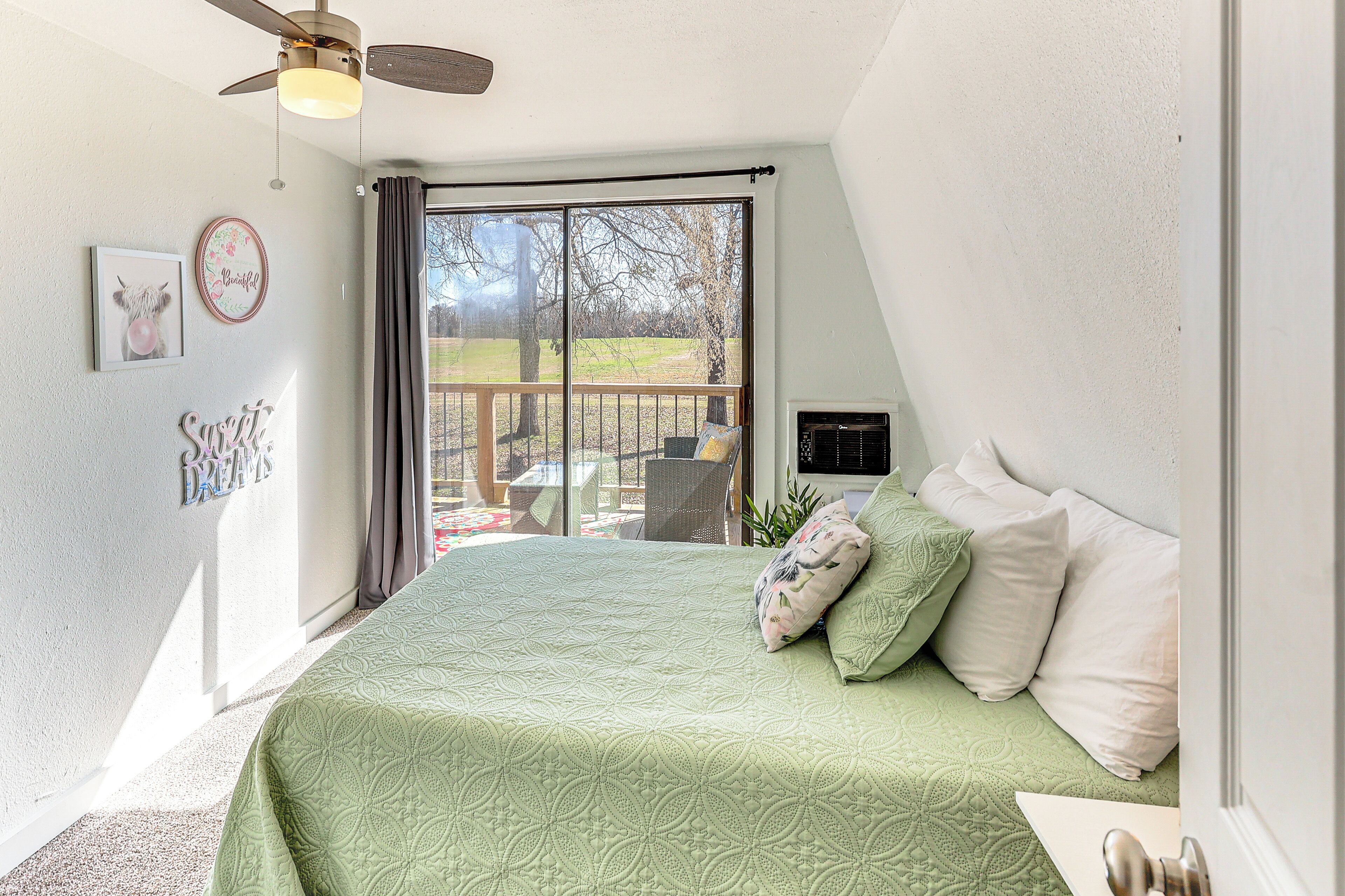 A bedroom with a green quilted bedspread, a ceiling fan, a large window with a view of a deck, and a door to the right.