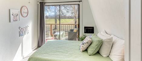 A bedroom with a green quilted bedspread, a ceiling fan, a large window with a view of a deck, and a door to the right.
