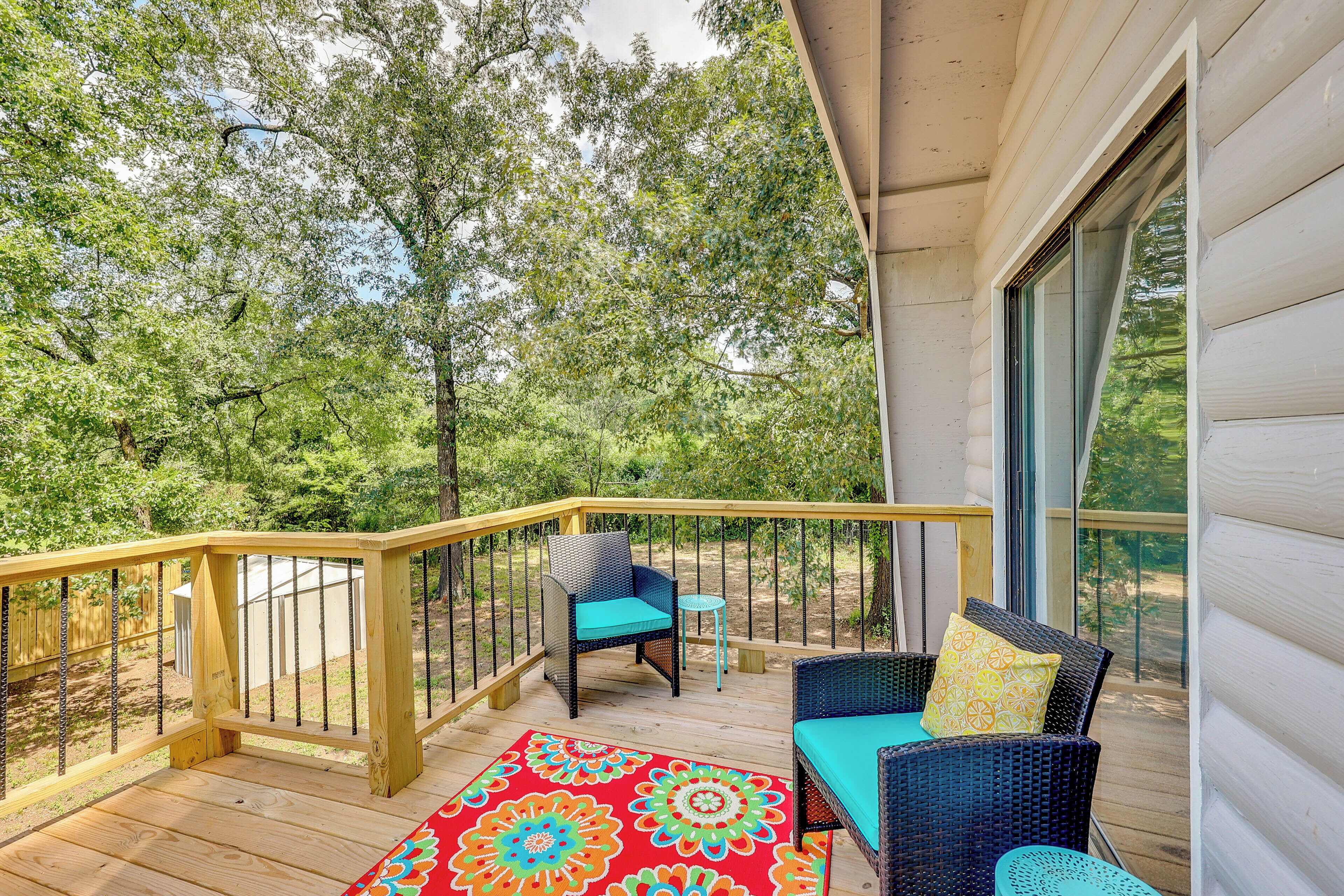 Une terrasse en bois, meublée de meubles en osier et agrémentée d ’un tapis coloré, le tout entouré d’arbres.