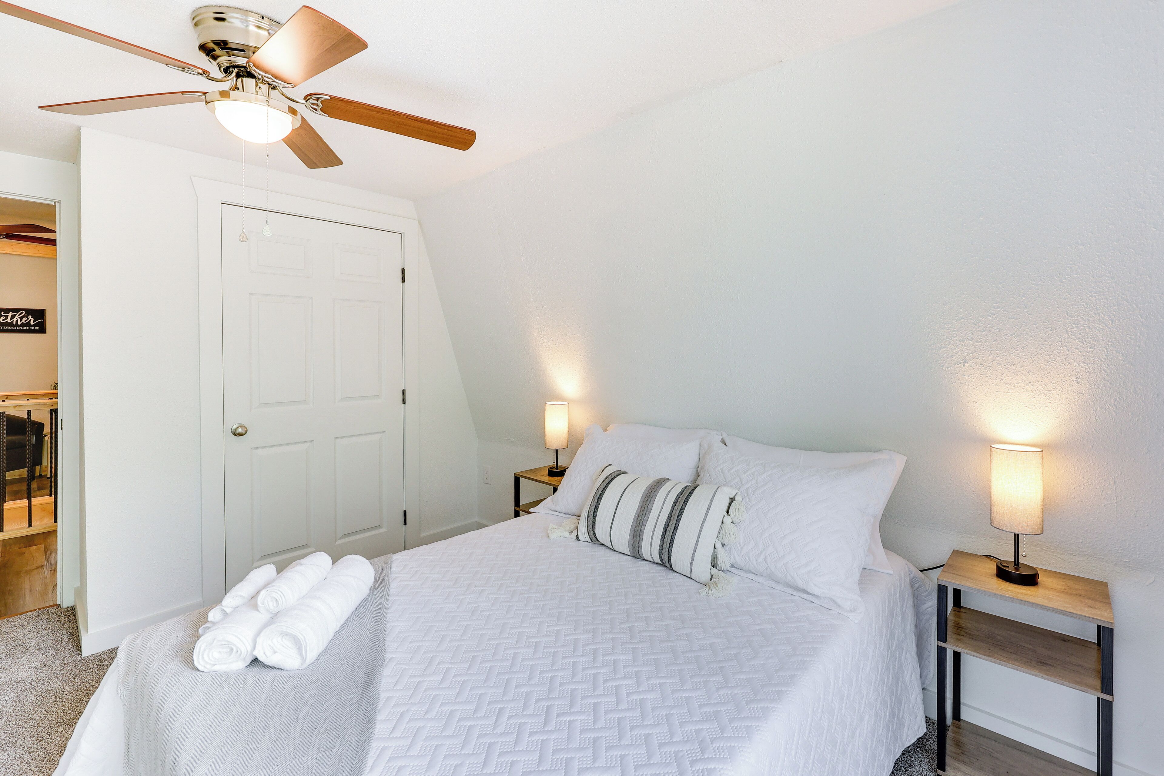 Un lit bien fait, avec du linge de lit blanc et un coussin à rayures, deux lampes de chevet, un ventilateur de plafond et une porte blanche.