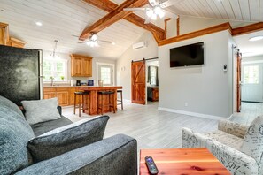 A spacious living area with a high ceiling, wooden beams, a kitchen with a bar, and a TV mounted on the wall.