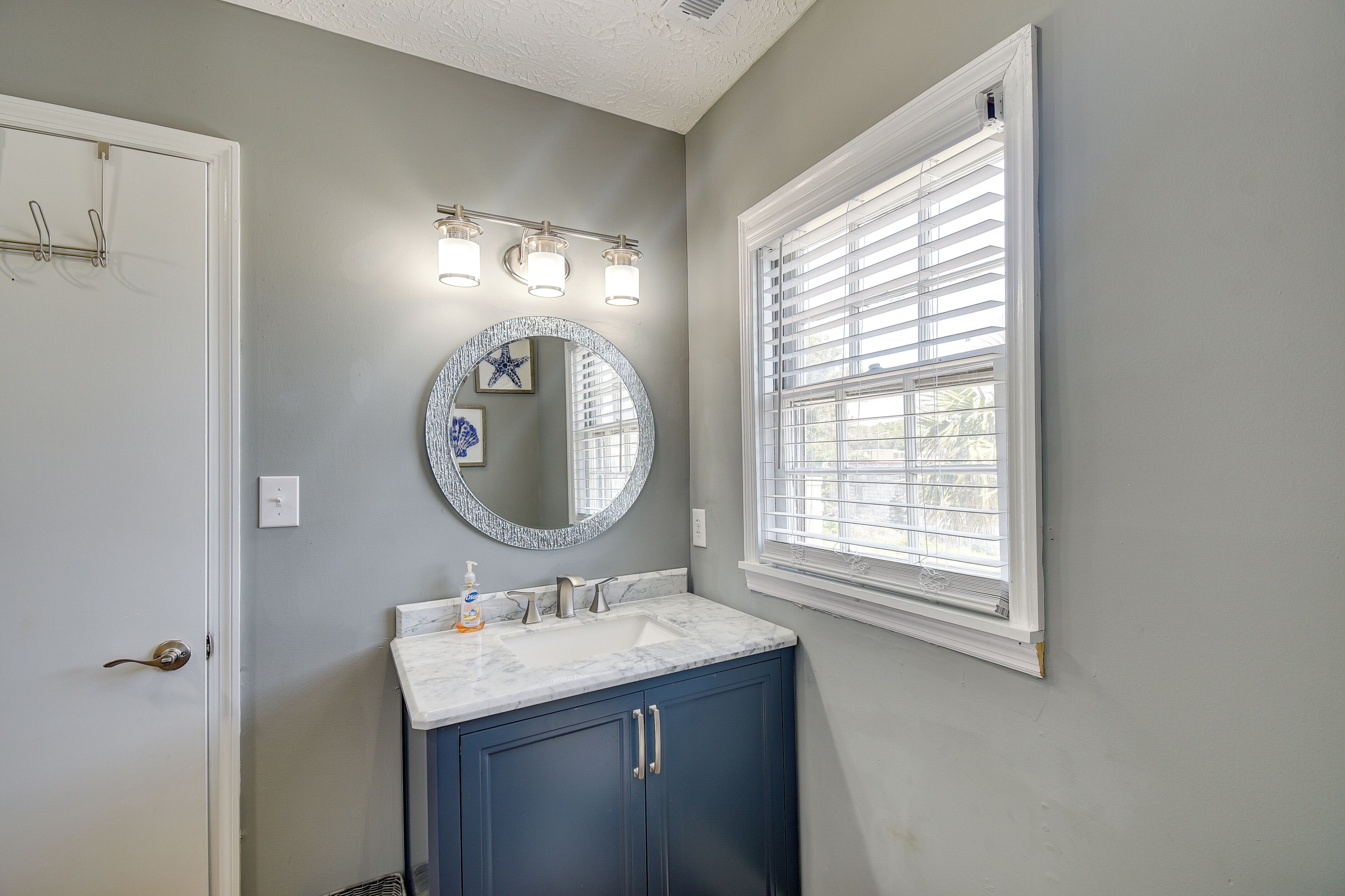 A bathroom with a vanity, a round mirror, and a window with blinds.