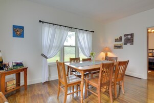 Un comedor con una mesa de madera, seis sillas y una ventana con cortinas blancas. Hay lámparas fijadas a la pared y cuadros enmarcados.