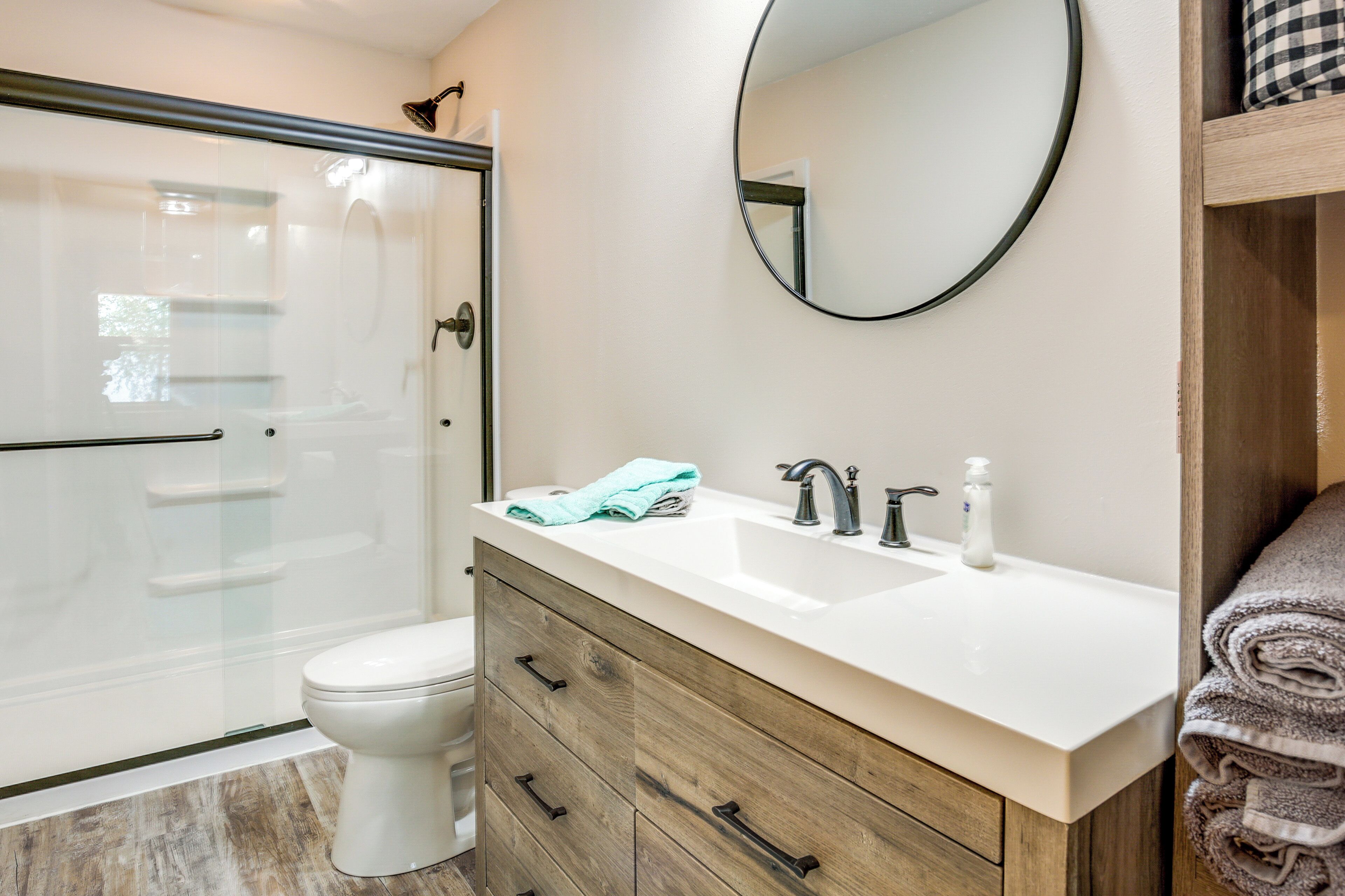 Une salle de bain avec une douche à parois de verre, des toilettes blanches, un meuble-lavabo en bois avec un lavabo blanc et un miroir rond.