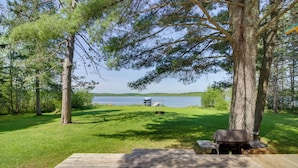 Una terraza de madera con una parrilla y una mesa, con vistas a un lago donde se divisa un barco a lo lejos, rodeada de árboles.