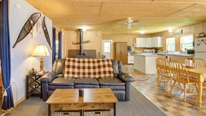 Una sala de estar acogedora con un sofá de cuero, una mesa de centro de madera y una manta de cuadros. Hay una zona de comedor con sillas de madera y una mesa, y una cocina con armarios blancos y una isla grande.