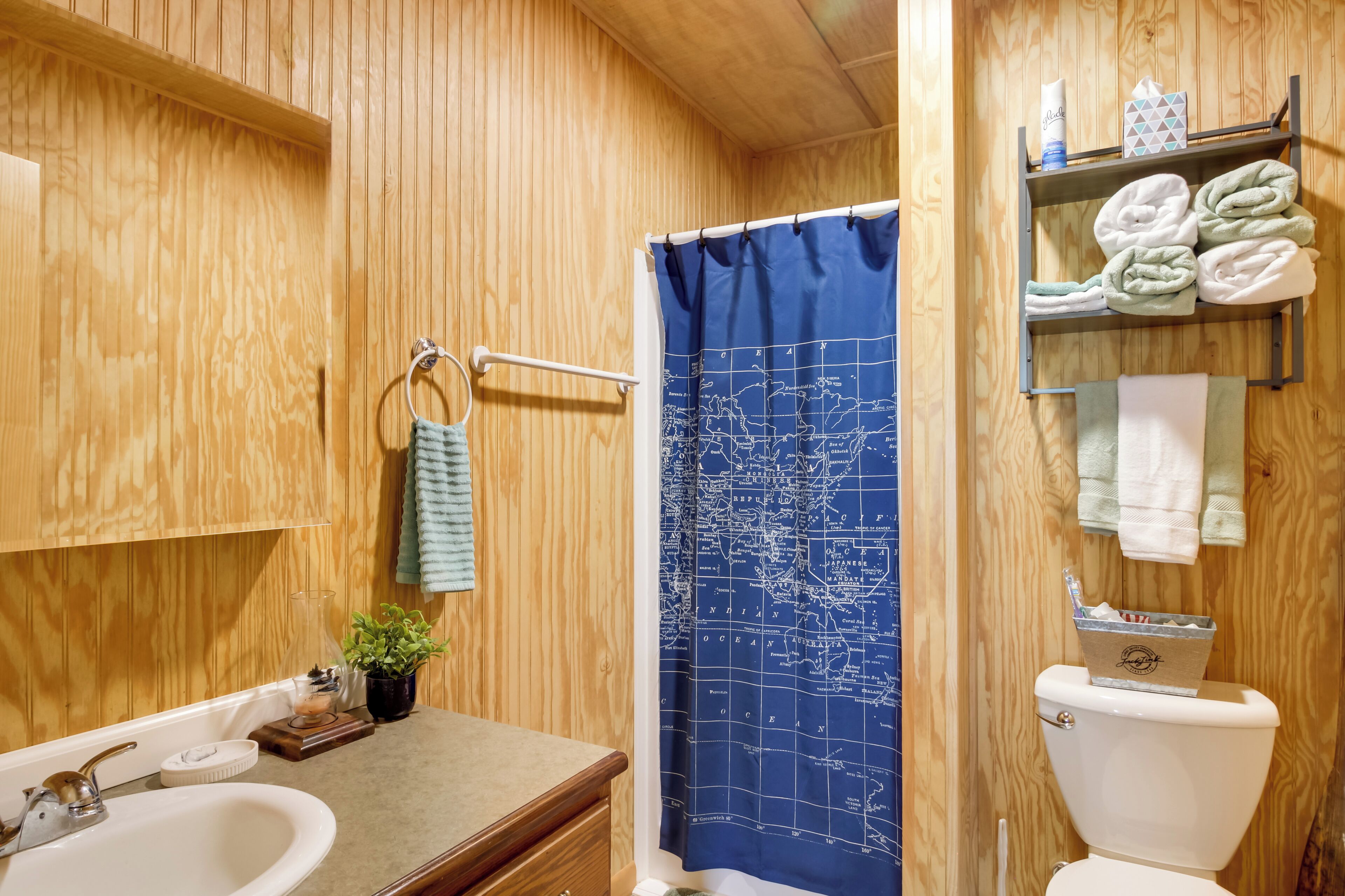 A wooden-paneled bathroom with a blue map shower curtain, a white sink, and a toilet with towels on a shelf.